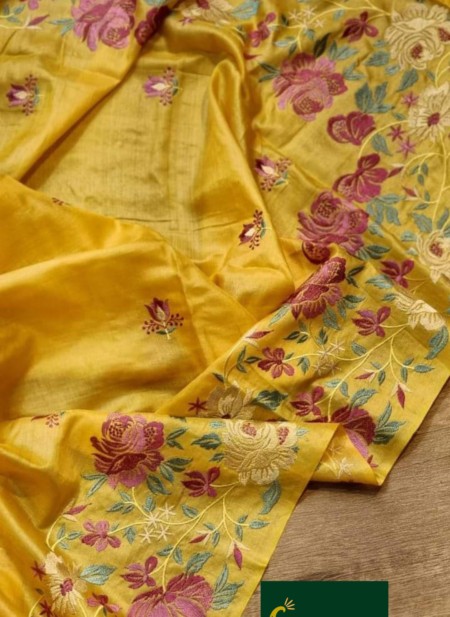A close-up shot of a mustard yellow Bhagalpuri Tussar silk saree featuring intricate pink, purple, and cream floral embroidery with colorful tassels.