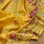 A close-up shot of a mustard yellow Bhagalpuri Tussar silk saree featuring intricate pink, purple, and cream floral embroidery with colorful tassels.