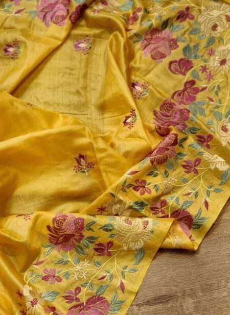 A close-up shot of a mustard yellow Bhagalpuri Tussar silk saree featuring intricate pink, purple, and cream floral embroidery with colorful tassels.