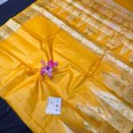 A mustard yellow Tussar silk Bhagalpuri saree with a broad gold zari border, featuring floral Meenakari work in pink and green, laid out on a dark surface.