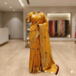 A mustard yellow Kota silk Bhagalpuri saree with red and green floral embroidery, gold zari borders, and maroon pompom edging on a mannequin.