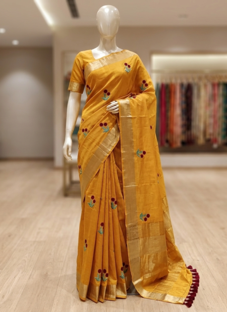 A mustard yellow Kota silk Bhagalpuri saree with red and green floral embroidery, gold zari borders, and maroon pompom edging on a mannequin.