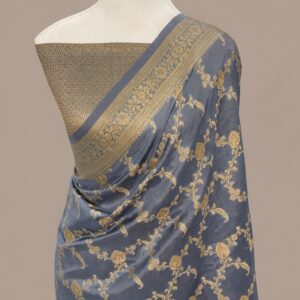 A Steel Grey Banarasi Soft Silk Saree featuring an intricate all-over antique gold zari floral jaal pattern, draped on a mannequin with a coordinating gold brocade blouse and a detailed traditional zari border.
