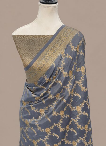A Steel Grey Banarasi Soft Silk Saree featuring an intricate all-over antique gold zari floral jaal pattern, draped on a mannequin with a coordinating gold brocade blouse and a detailed traditional zari border.