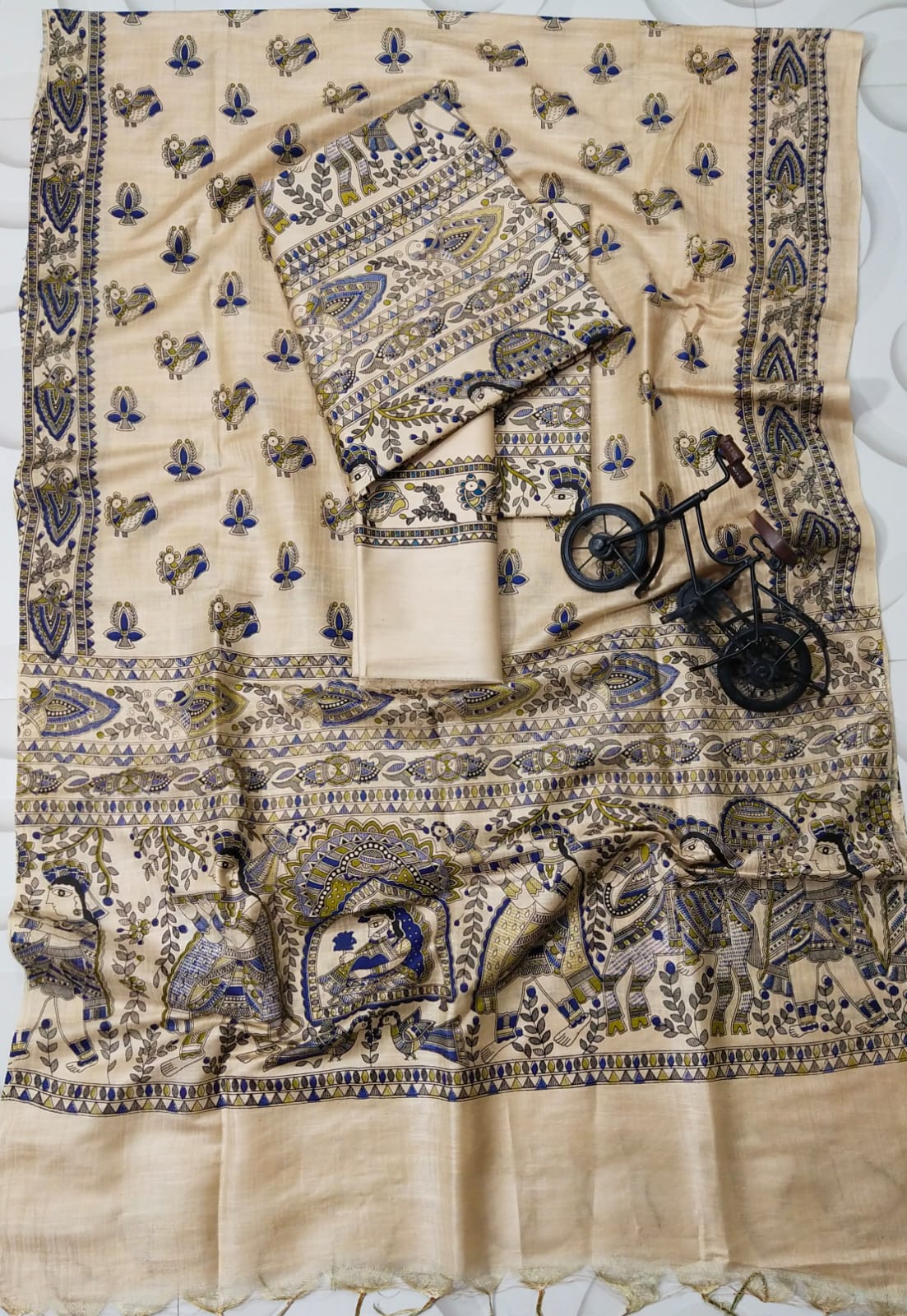 Beige & Indigo Blue Madhubani Print Unstitched Katan Silk Suit Material by Bhavya Lakshmi Fabrics An unstitched cream Katan silk suit material set by Bhavya Lakshmi Fabrics featuring detailed multi-colored Madhubani prints of ceremonial figures, elephants, and peacocks in indigo blue and olive green.
