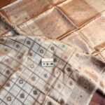 Natural beige pure Bhagalpuri Munga silk saree from Bhavya Lakshmi Fabrics featuring gold zari checks with multi-color floral Meenakari butis and an intricate gold zari border, spread on a wooden floor.