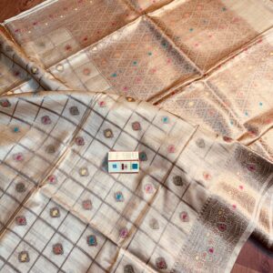 Natural beige pure Bhagalpuri Munga silk saree from Bhavya Lakshmi Fabrics featuring gold zari checks with multi-color floral Meenakari butis and an intricate gold zari border, spread on a wooden floor.