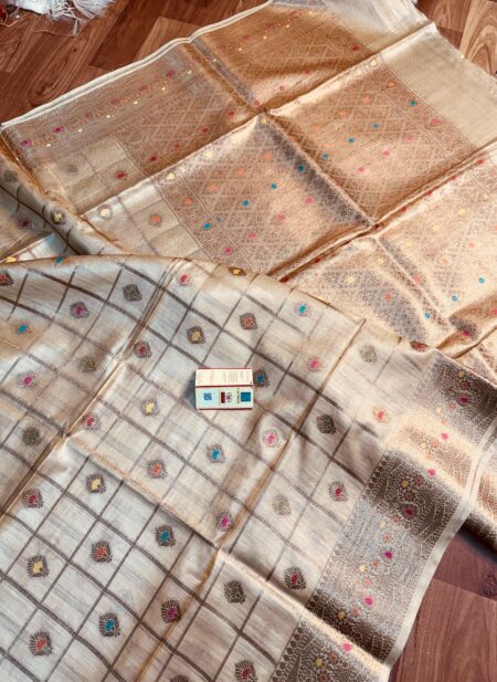 Natural beige pure Bhagalpuri Munga silk saree from Bhavya Lakshmi Fabrics featuring gold zari checks with multi-color floral Meenakari butis and an intricate gold zari border, spread on a wooden floor.