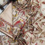 A premium unstitched beige Katan silk suit material set by Bhavya Lakshmi Fabrics featuring detailed multicolored Madhubani art prints of fluting deities, birds, and fish on a clean, minimal off-white backdrop.