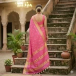 A woman wearing a vibrant pink Kota Silk Bhagalpuri saree with a wide gold zari border and white tassels standing on stone steps.