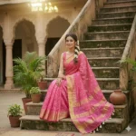 A woman wearing a vibrant pink Kota Silk Bhagalpuri saree with a wide gold zari border and white tassels standing on stone steps.