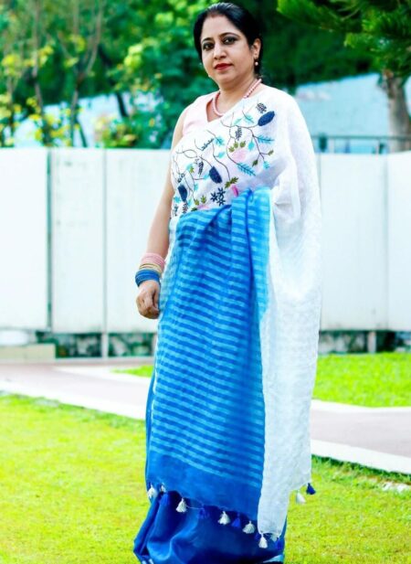 An Indian model showcases a white pure linen saree by Bhavya Lakshmi Fabrics featuring vibrant blue and pink floral embroidery and a gradient blue border.