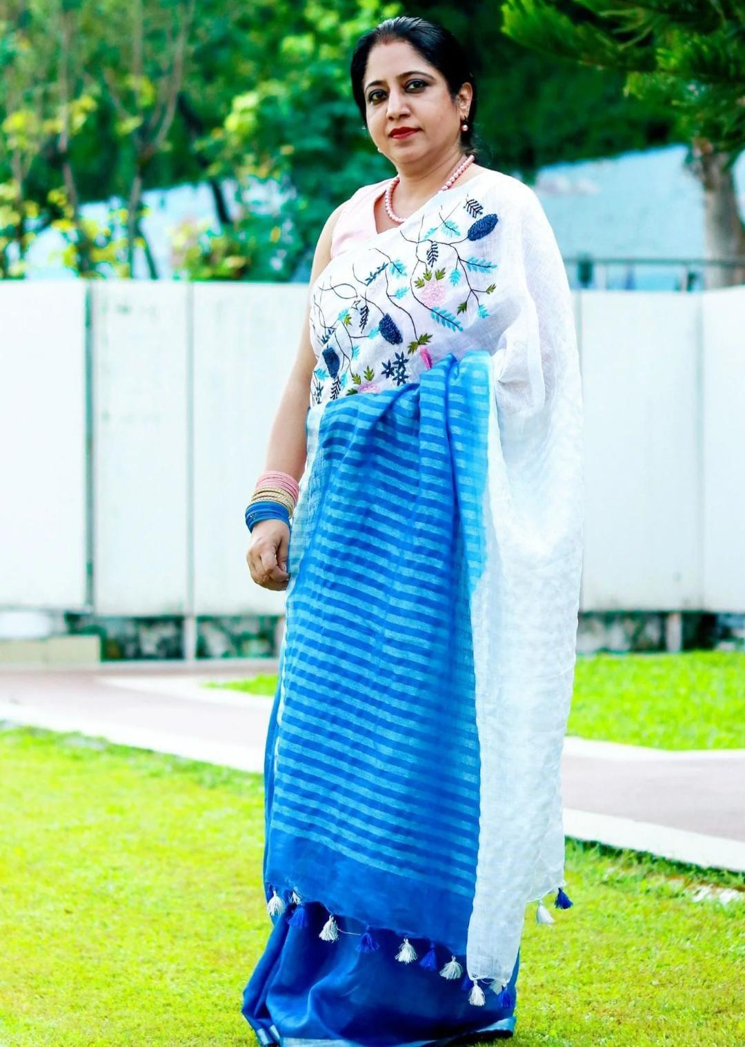 White and Blue Embroidered Bhagalpuri Pure Linen Saree by Bhavya Lakshmi Fabrics An Indian model showcases a white pure linen saree by Bhavya Lakshmi Fabrics featuring vibrant blue and pink floral embroidery and a gradient blue border.