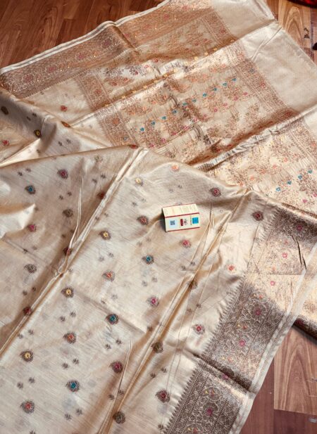 Natural beige Pure Bhagalpuri Muga silk saree featuring small multi-color floral butis, an intricate zari border, and a heavily woven floral pallu spread on a wooden floor.
