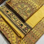 An unstitched mustard yellow Katan silk suit material set by Bhavya Lakshmi Fabrics featuring extensive multi-colored Madhubani prints of ceremonial elephants, ritual huts, and stylized birds with geometric circular borders.