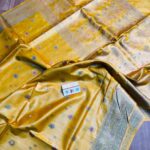 Alt Text:Vibrant mustard yellow pure Bhagalpuri Munga silk saree with small multi-color meenakari butis and an intricate zari border, spread on a wooden floor.