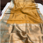 A close-up view of a sheer Mustard Yellow Organza Banarasi Saree from Bhavya Lakshmi Fabrics featuring detailed handwoven silver zari butis and a wide lattice-patterned silver border.