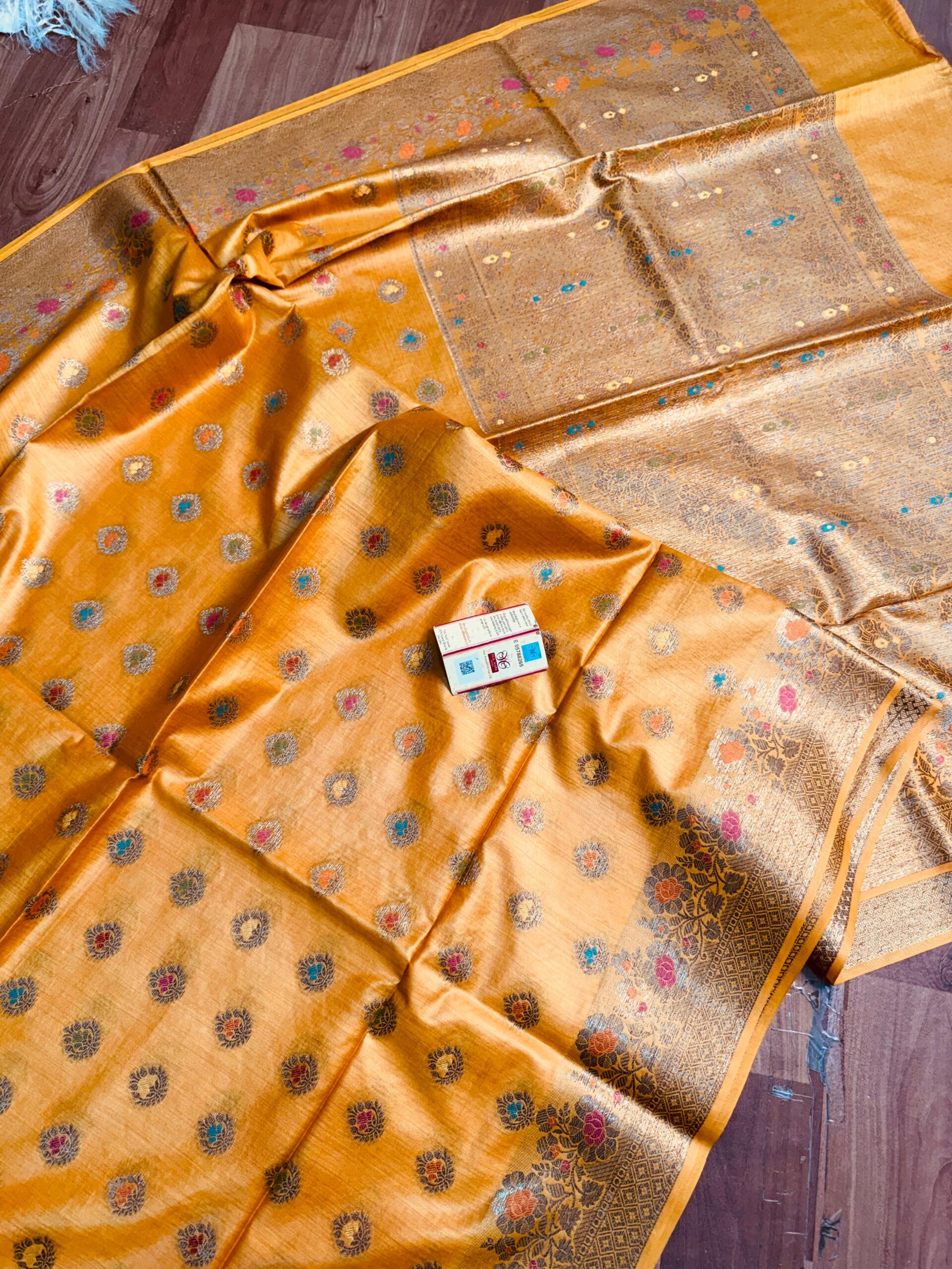 Mustard Yellow Pure Bhagalpuri Munga Silk Saree by Bhavya Lakshmi Fabrics (3) Vibrant mustard yellow pure Bhagalpuri Munga silk saree from Bhavya Lakshmi Fabrics featuring small multi-color Meenakari floral butis and an intricate gold zari border, spread on a wooden floor.