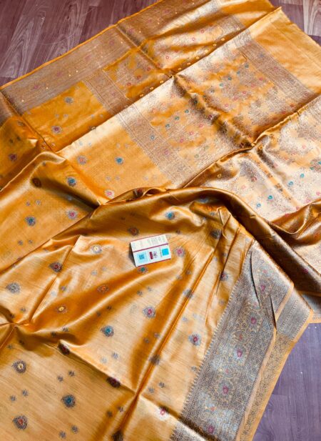 Vibrant mustard yellow pure Bhagalpuri Munga silk saree from Bhavya Lakshmi Fabrics featuring small multi-color Meenakari floral butis and an intricate gold zari border, spread on a wooden floor.