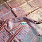 Elegant pastel peach pure Bhagalpuri Munga silk saree from Bhavya Lakshmi Fabrics featuring gold zari checks with multi-color floral Meenakari butis and an intricate gold zari border, spread on a wooden floor.