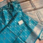 Vibrant peacock blue pure Bhagalpuri Munga silk saree from Bhavya Lakshmi Fabrics featuring gold zari checks with multi-color floral Meenakari butis and an intricate gold zari border, spread on a wooden floor.