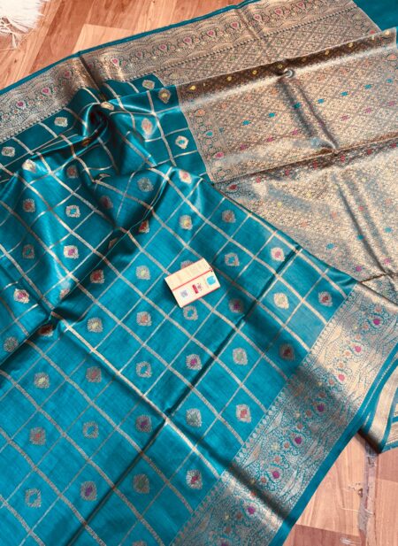 Vibrant peacock blue pure Bhagalpuri Munga silk saree from Bhavya Lakshmi Fabrics featuring gold zari checks with multi-color floral Meenakari butis and an intricate gold zari border, spread on a wooden floor.