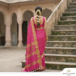 A woman wearing a vibrant pink Kota Silk Bhagalpuri saree with a wide gold zari border and white tassels standing on stone steps.