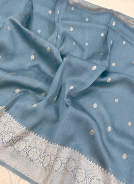 A close-up view of a serene Powder Blue Organza Banarasi Saree from Bhavya Lakshmi Fabrics featuring detailed handwoven silver zari floral borders and delicate scattered buti