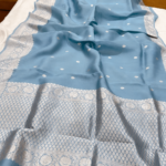 A close-up view of a serene Powder Blue Organza Banarasi Saree from Bhavya Lakshmi Fabrics featuring detailed handwoven silver zari floral borders and delicate scattered butis