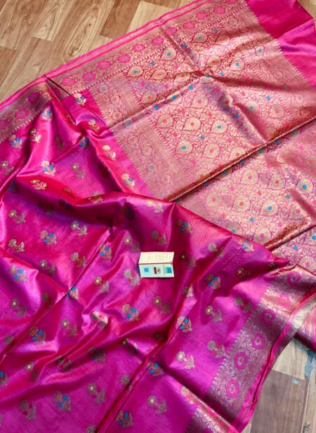 Vibrant Rani pink pure Bhagalpuri Munga silk saree from Bhavya Lakshmi Fabrics featuring multi-color floral butis and an intricate gold zari border, spread on a wooden floor.