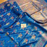 Vibrant royal blue pure Bhagalpuri Munga silk saree from Bhavya Lakshmi Fabrics featuring a multi-color floral Meenakari jaal and an intricate gold zari border, spread on a wooden floor