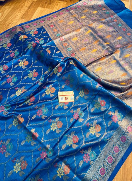 Vibrant royal blue pure Bhagalpuri Munga silk saree from Bhavya Lakshmi Fabrics featuring a multi-color floral Meenakari jaal and an intricate gold zari border, spread on a wooden floor