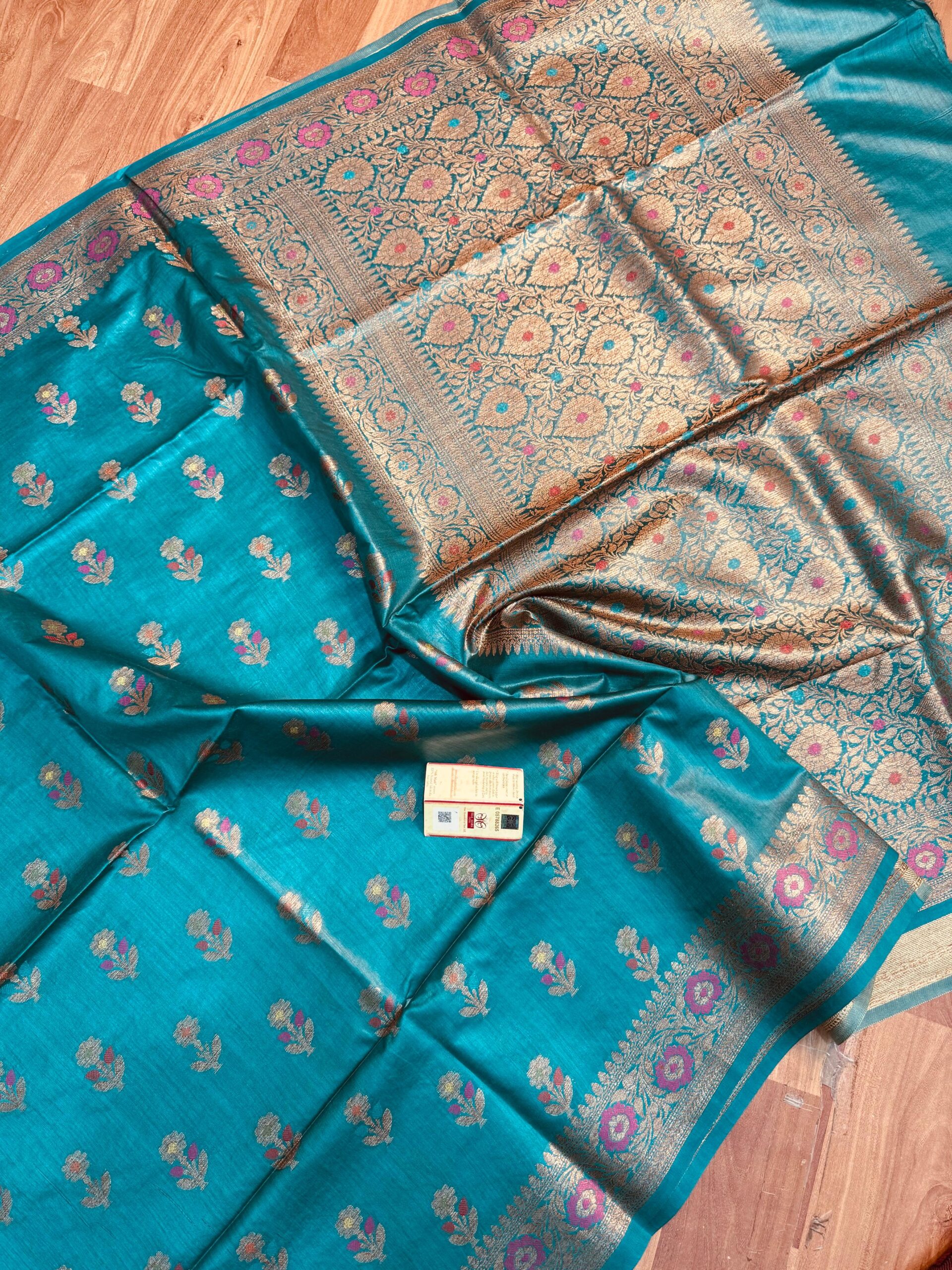 Sea Green Pure Bhagalpuri Munga Silk Saree by Bhavya Lakshmi Fabrics Vibrant sea green pure Bhagalpuri Munga silk saree from Bhavya Lakshmi Fabrics featuring small multi-color floral butis and an intricate gold zari border with pink accents, spread on a wooden floor.