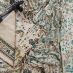 An unstitched beige Katan silk suit material set by Bhavya Lakshmi Fabrics featuring detailed teal blue and mustard yellow Warli tribal dance prints, geometric borders, and a matching dupatta