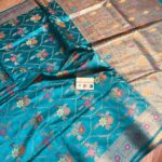 Vibrant turquoise blue pure Bhagalpuri Munga silk saree from Bhavya Lakshmi Fabrics featuring a multi-color floral Meenakari jaal and an intricate gold zari border, spread on a wooden floor.