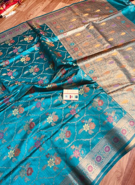 Vibrant turquoise blue pure Bhagalpuri Munga silk saree from Bhavya Lakshmi Fabrics featuring a multi-color floral Meenakari jaal and an intricate gold zari border, spread on a wooden floor.