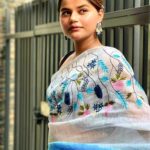 An Indian model showcases a white pure linen saree by Bhavya Lakshmi Fabrics featuring vibrant blue and pink floral embroidery and a gradient blue border.