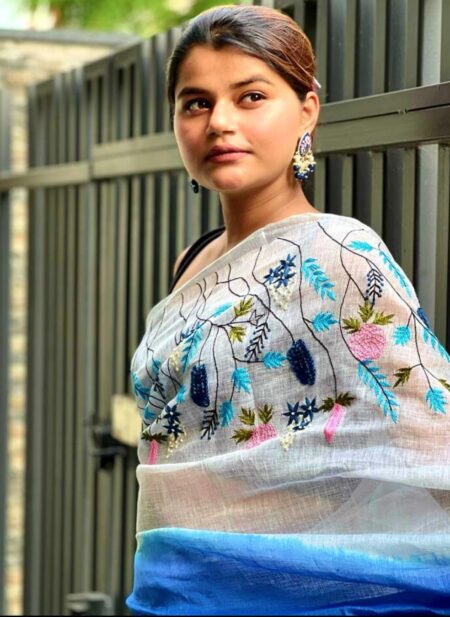 An Indian model showcases a white pure linen saree by Bhavya Lakshmi Fabrics featuring vibrant blue and pink floral embroidery and a gradient blue border.