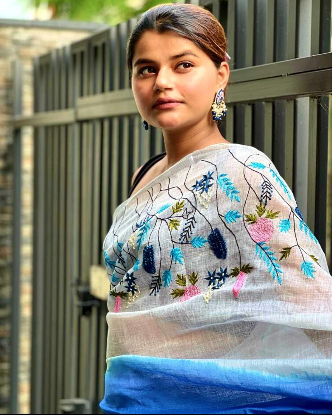 White and Blue Embroidered Bhagalpuri Pure Linen Saree by Bhavya Lakshmi Fabrics An Indian model showcases a white pure linen saree by Bhavya Lakshmi Fabrics featuring vibrant blue and pink floral embroidery and a gradient blue border.