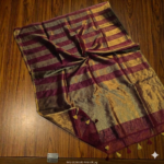 Rich maroon and metallic gold zari striped tissue silk saree featuring intricate borders and matching tassel details.