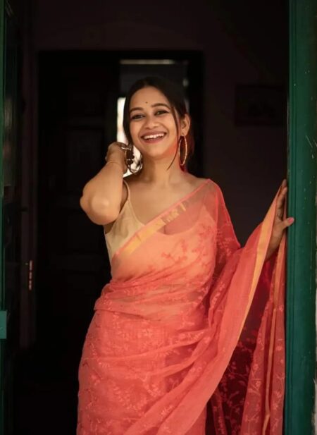 Bhavyaloom peach pink bhagalpuri kota doria embroidery saree with gold border on smiling model standing in doorway