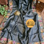 "A luxurious charcoal dark grey pure Tussar Munga silk saree laid flat, featuring scattered small dark embroidered dots across the body and an intricate border adorned with vibrant multicolored floral embroidery in pink, peach, violet, and green tones. An antique brass incense burner sits elegantly on top."