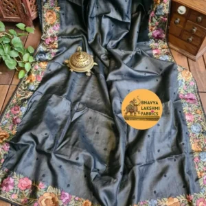 "A luxurious charcoal dark grey pure Tussar Munga silk saree laid flat, featuring scattered small dark embroidered dots across the body and an intricate border adorned with vibrant multicolored floral embroidery in pink, peach, violet, and green tones. An antique brass incense burner sits elegantly on top."