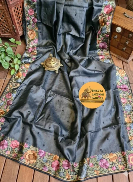 "A luxurious charcoal dark grey pure Tussar Munga silk saree laid flat, featuring scattered small dark embroidered dots across the body and an intricate border adorned with vibrant multicolored floral embroidery in pink, peach, violet, and green tones. An antique brass incense burner sits elegantly on top."