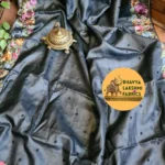 "A luxurious charcoal dark grey pure Tussar Munga silk saree laid flat, featuring scattered small dark embroidered dots across the body and an intricate border adorned with vibrant multicolored floral embroidery in pink, peach, violet, and green tones. An antique brass incense burner sits elegantly on top."