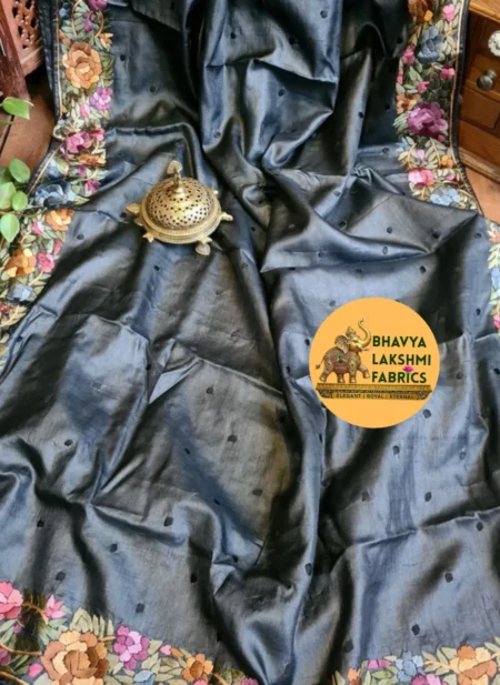 "A luxurious charcoal dark grey pure Tussar Munga silk saree laid flat, featuring scattered small dark embroidered dots across the body and an intricate border adorned with vibrant multicolored floral embroidery in pink, peach, violet, and green tones. An antique brass incense burner sits elegantly on top."