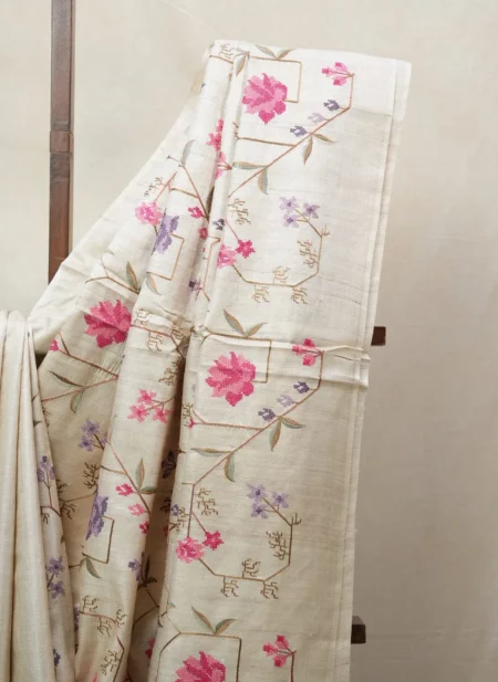 Bhavyaloom natural cream 100 percent pure tussar muga silk saree with pink and purple floral hand embroidery draped on wooden stand