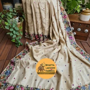 A luxurious natural cream beige pure Tussar Munga silk saree draped elegantly on a wooden floor background, featuring scattered small colorful embroidered dots across the body and an intricate border adorned with vibrant multicolored floral embroidery in shades of magenta, purple, blue, and peach with green leaves.