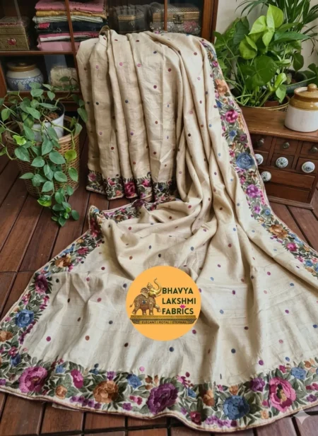 A luxurious natural cream beige pure Tussar Munga silk saree draped elegantly on a wooden floor background, featuring scattered small colorful embroidered dots across the body and an intricate border adorned with vibrant multicolored floral embroidery in shades of magenta, purple, blue, and peach with green leaves.