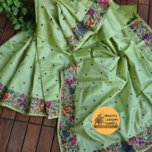 "A luxurious pista green pure Tussar Munga silk saree laid flat, featuring scattered dark embroidered dots across the body and an intricate border adorned with vibrant multicolored floral embroidery in shades of magenta, violet, indigo, and orange with green leaves. The textile displays a rich, textured wild silk grain